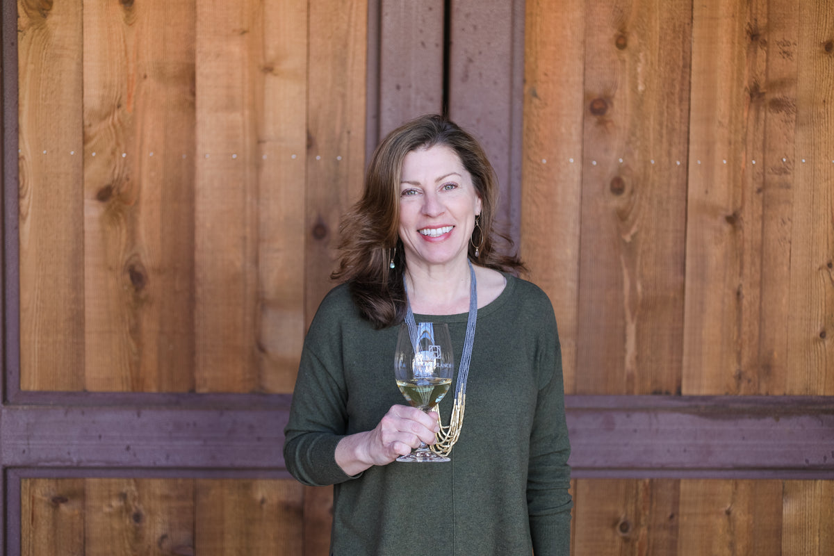 A smiling woman with brown hair stands in front of a wooden wall, holding a glass of white wine. She is wearing a dark olive green long-sleeve top and a gold necklace.