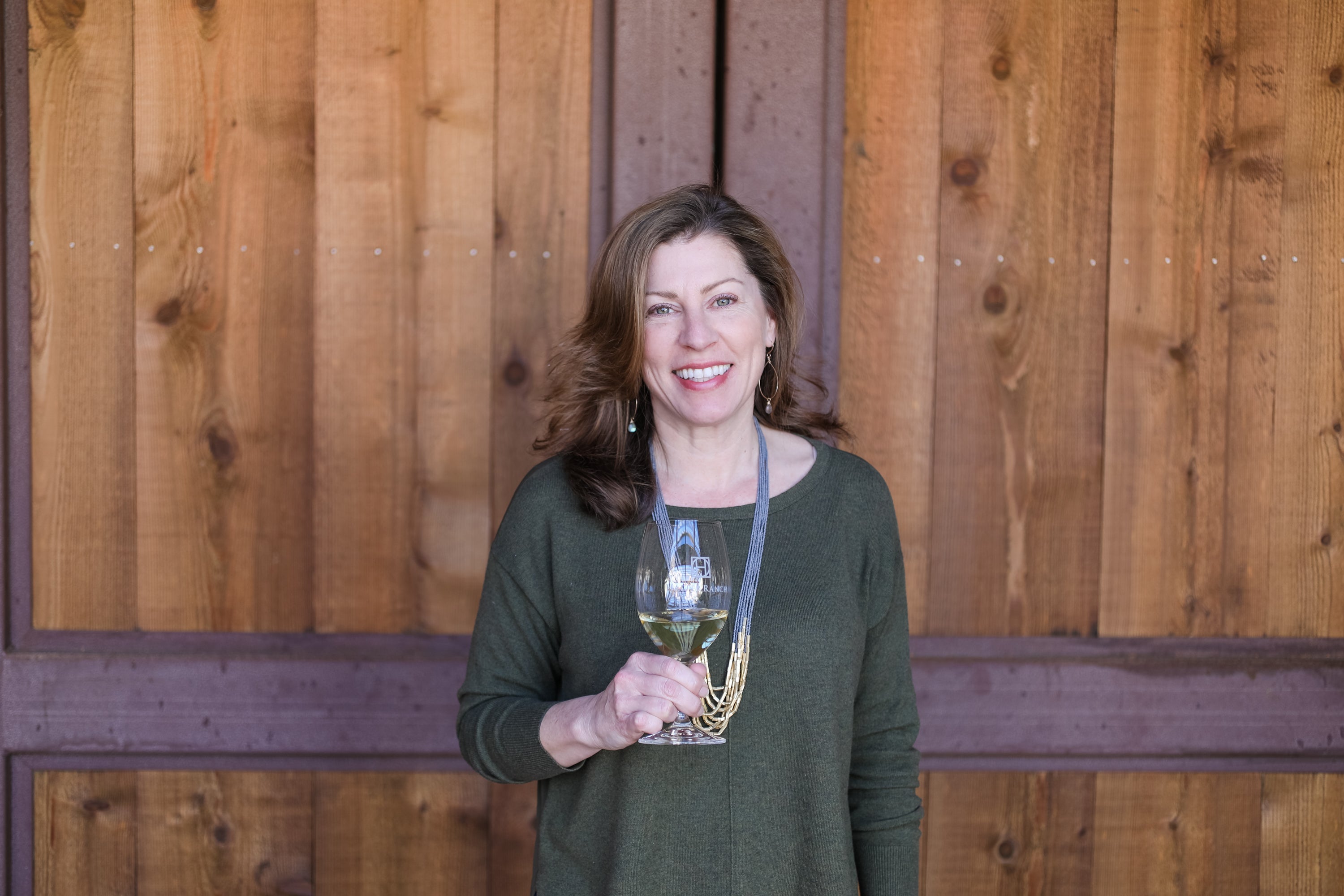 A smiling woman with brown hair stands in front of a wooden wall, holding a glass of white wine. She is wearing a dark olive green long-sleeve top and a gold necklace.